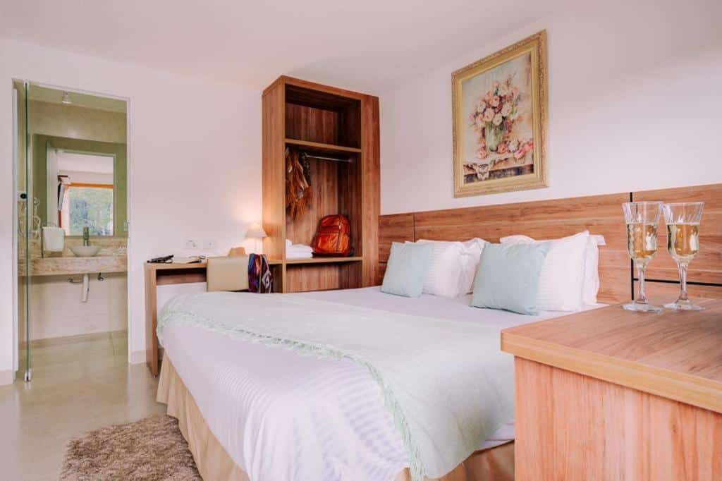 Uma cama bem arrumada em um quarto do Hotel Portal das Videiras com móveis de madeira, uma escrivaninha e duas taças de champanhe na mesa de cabeceira, para representar hotéis em Monte Verde.