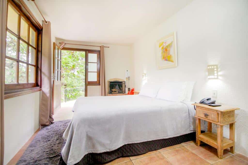 Um quarto bem iluminado no VELINN Hotel Ninho do Falcão com uma cama grande, mesa de cabeceira de madeira, lareira e portas abertas que levam a uma varanda com vegetação externa, para representar hotéis em Monte Verde.