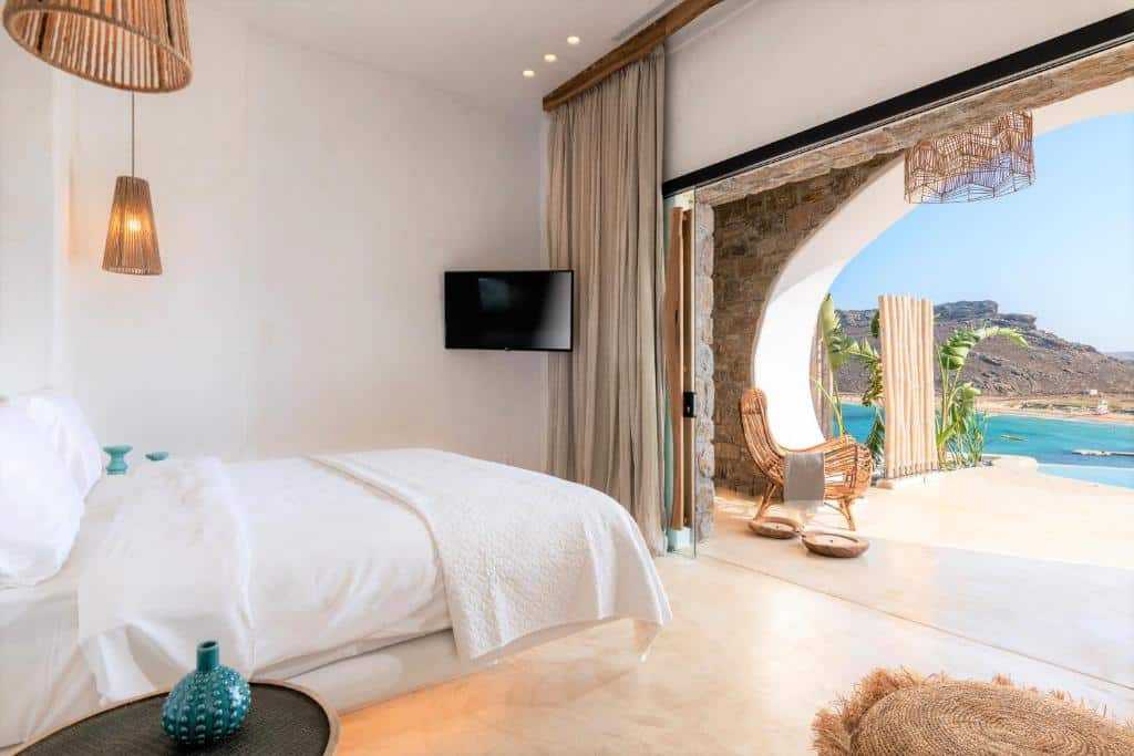 Um quarto no Panormos Village bem iluminado com uma cama, TV e portas abertas que levam a uma varanda com vista para o oceano e a costa, para representar hotéis em Mykonos.