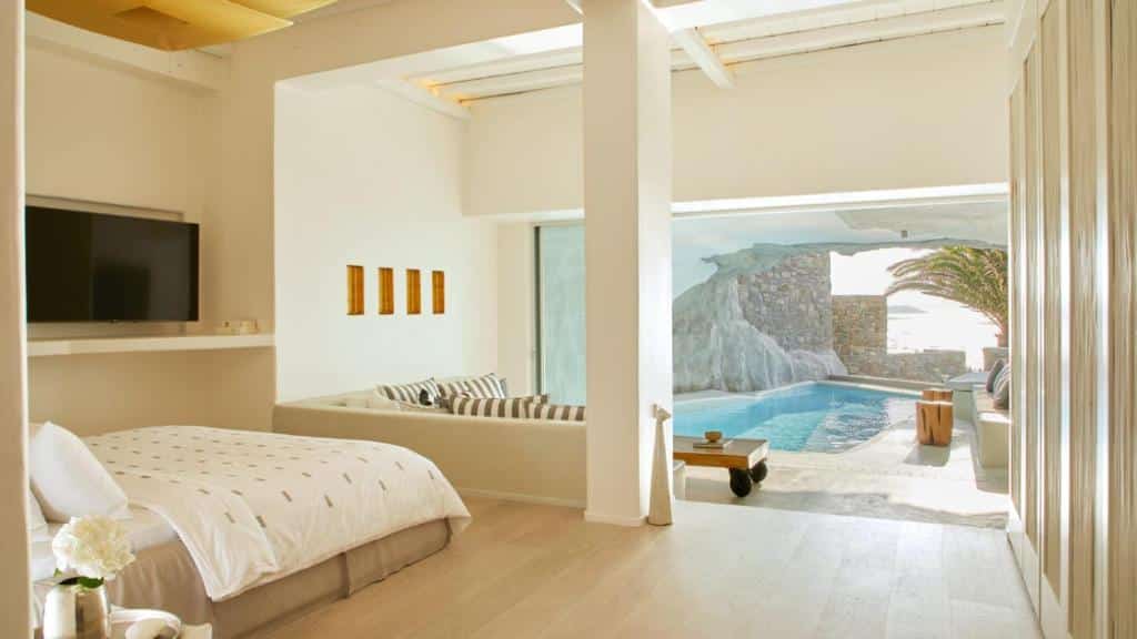 Um quarto no Cavo Tagoo Mykonos moderno com cama, sofá, TV e uma grande janela que dá para uma piscina privativa e uma área de pátio ensolarada, para representar hotéis em Mykonos.