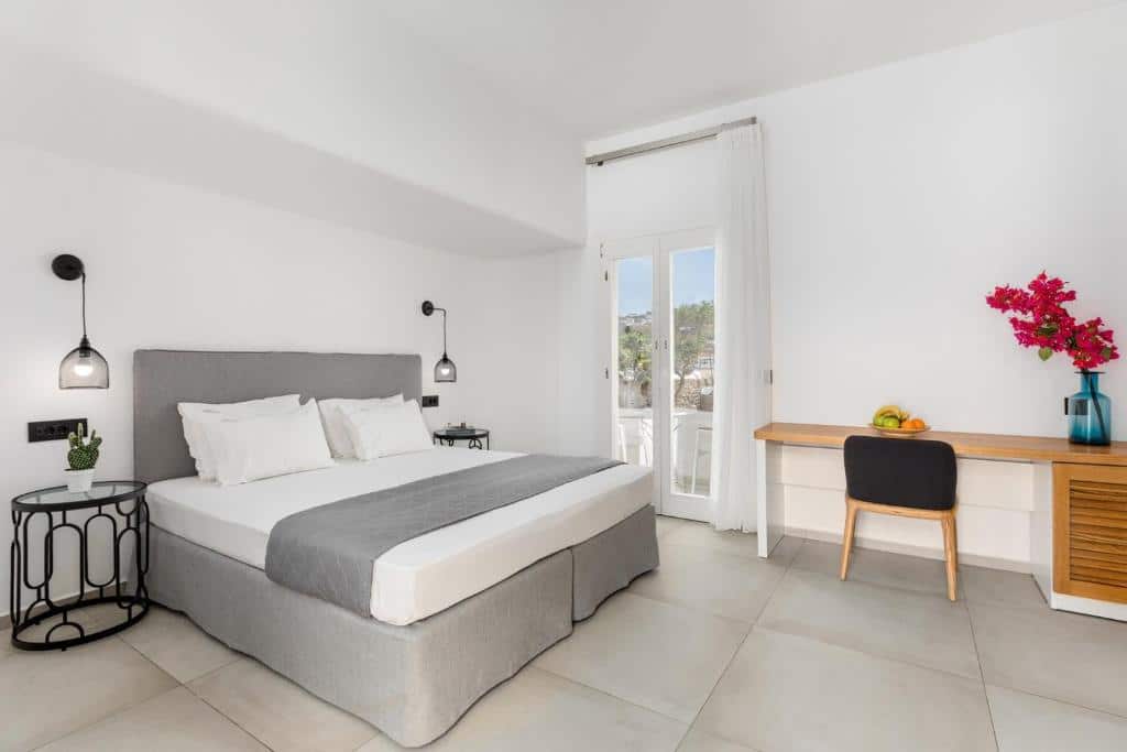 Um quarto moderno no Corfos Hotel com uma cama de casal, uma mesa lateral, uma escrivaninha com uma cadeira e um vaso com flores cor-de-rosa perto de uma janela, para representar hotéis em Mykonos.