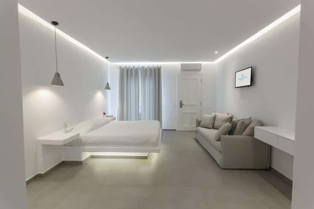 Quarto do Aeolos Resort branco minimalista com cama flutuante, sofá cinza, TV na parede, luzes pendentes e janela grande com cortinas transparentes, para representar hotéis em Mykonos.