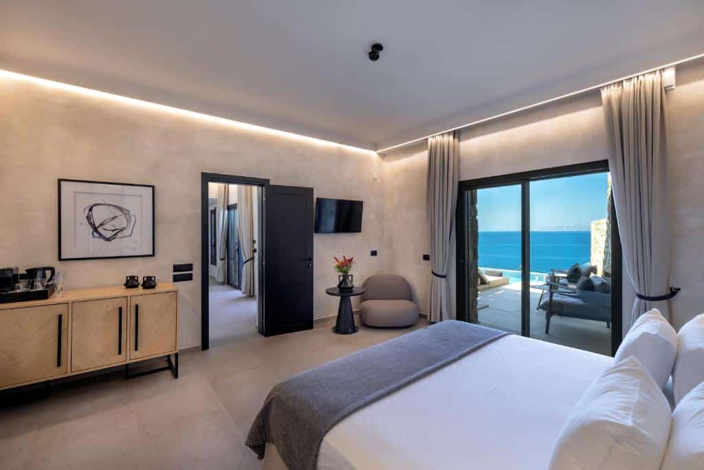 Quarto moderno no Tropicana Hotel, Suites & Villas Mykonos com uma cama grande, TV, aparador, cadeira e portas de varanda com vista para o oceano, para representar hotéis em Mykonos.