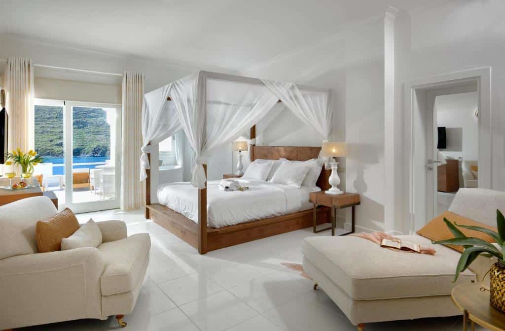 Quarto no Nissaki Boutique Hotel bem iluminado com cama de dossel, móveis brancos, livro aberto e varanda com vista para a água e as colinas, para representar hotéis em Mykonos.