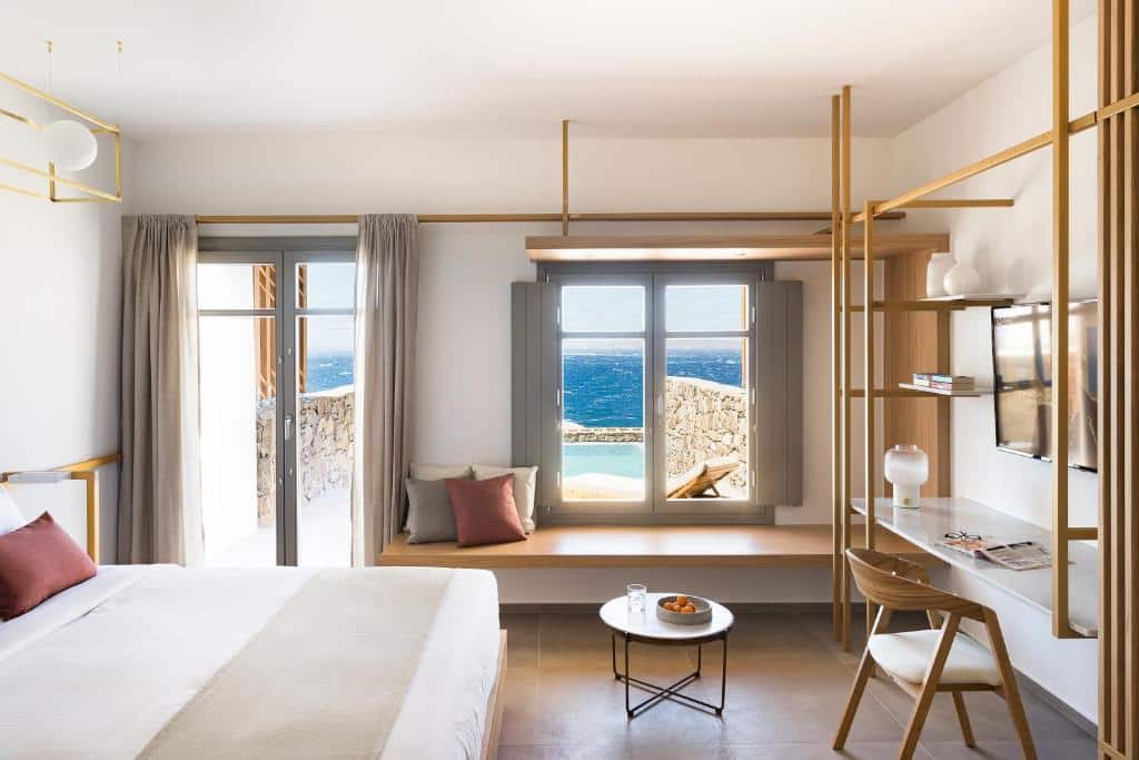 Um quarto moderno no Mykonos Soul Luxury Suites com cama, escrivaninha e assento na janela, com vista para o mar através de grandes janelas, para rperesentar hotéis em Mykonos.