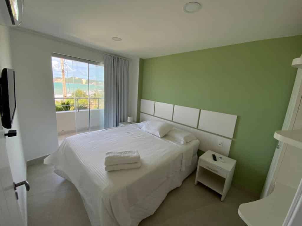 Uma cama bem arrumada com lençóis brancos em um quarto bem iluminado com parede verde, janela e vista para a varanda.