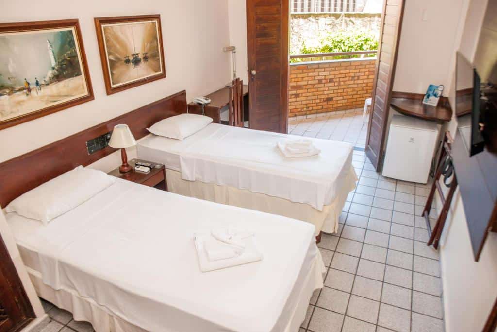 Quarto de hotel com duas camas de solteiro, roupa de cama branca, mesa de cabeceira, piso de azulejo, TV, frigobar e porta para o pátio externo.