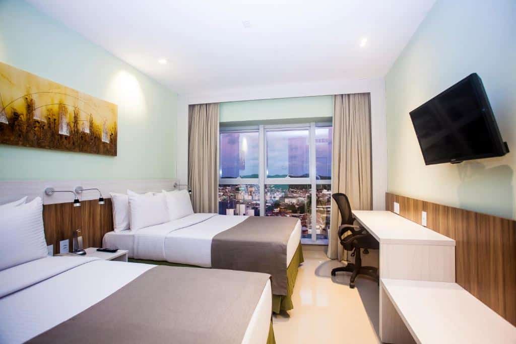 Quarto de hotel com duas camas de casal, uma TV na parede, escrivaninha com cadeira e uma janela com vista para a paisagem urbana.
