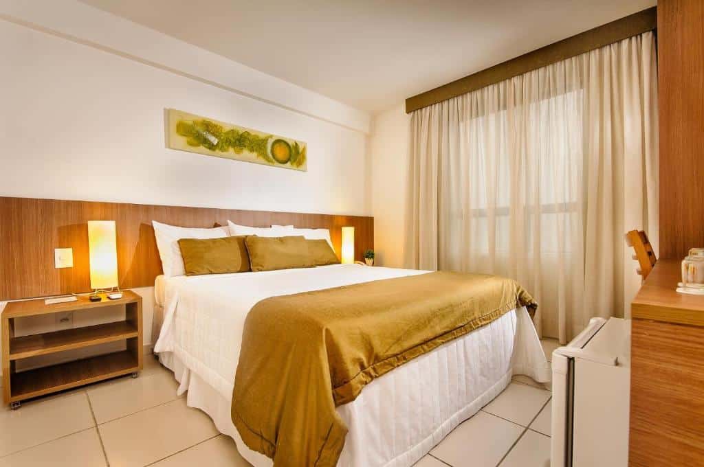 Um quarto de hotel com uma cama de casal, roupa de cama marrom, duas mesas de cabeceira com luminárias e uma janela com cortinas transparentes.