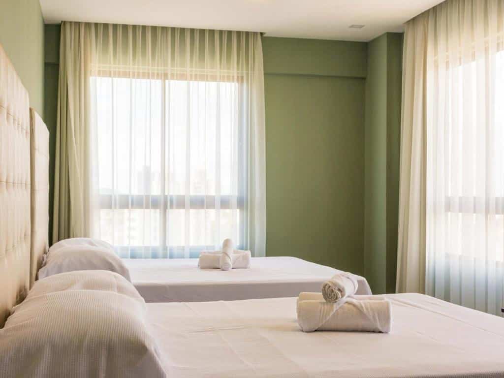 Duas camas bem arrumadas com lençóis brancos e toalhas enroladas em um quarto bem iluminado com janelas grandes e cortinas transparentes.