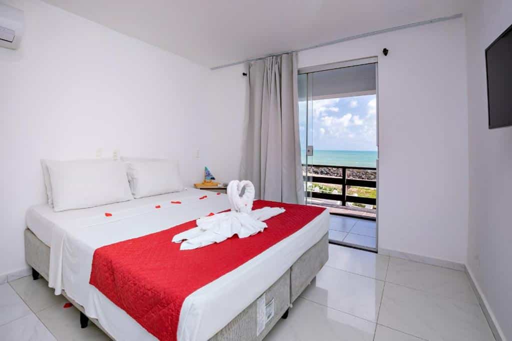 Um quarto de hotel com cama de casal, corredor vermelho, cisne de toalha, varanda e vista para o mar sob um céu parcialmente nublado.