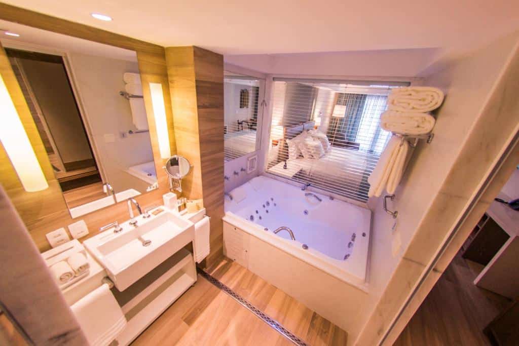 Banheiro de hotel moderno com uma banheira grande, pias duplas, espelho, toalhas e detalhes em madeira.