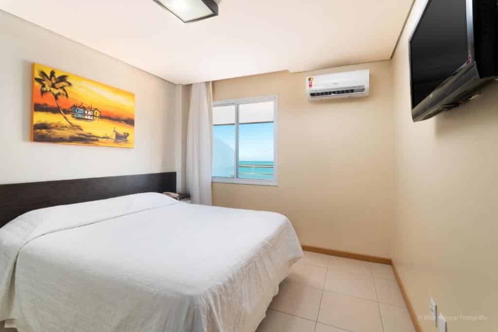 Um pequeno quarto de hotel com uma cama de casal, arte na parede, janela com vista para o mar, ar-condicionado e TV na parede.