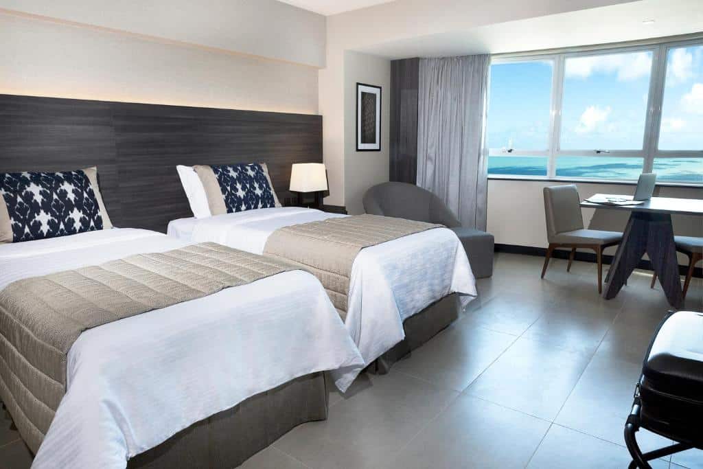 Quarto de hotel com duas camas, um abajur, uma poltrona, uma escrivaninha perto de uma janela e vista para o oceano do lado de fora.