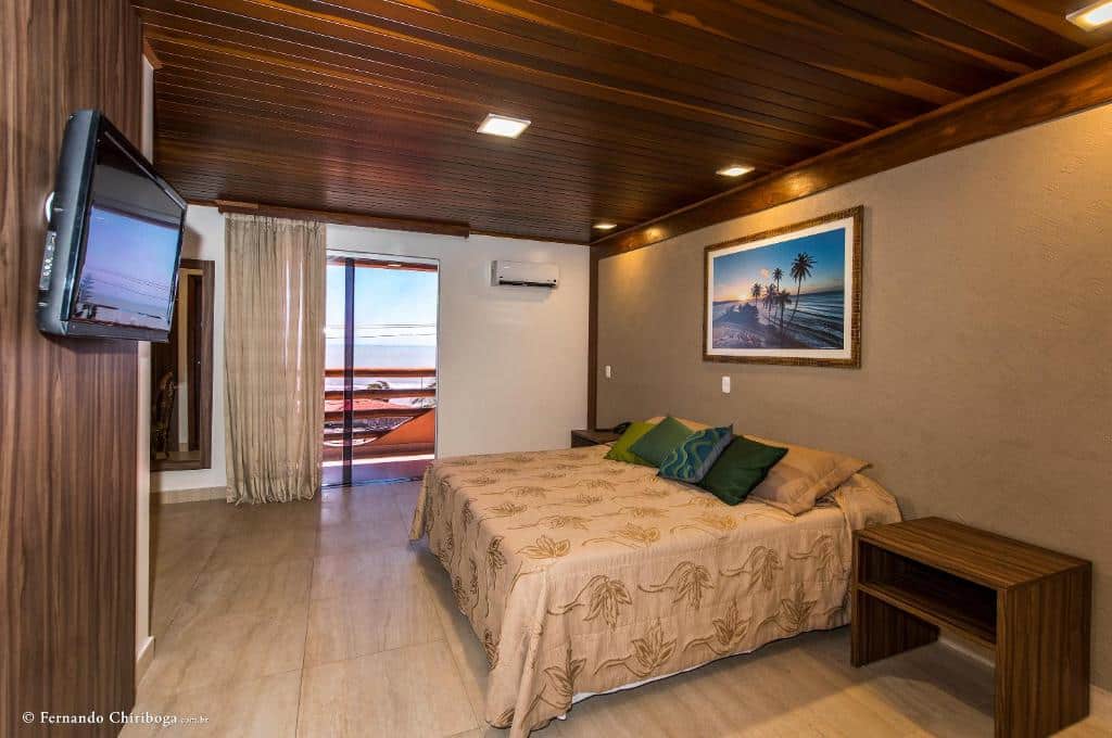 Um quarto de hotel com uma cama de casal, duas mesinhas de cabeceira, arte na parede, uma TV e uma varanda com vista para o oceano.