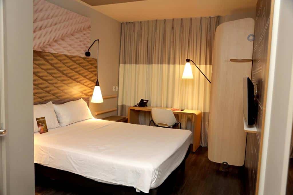 Quarto de hotel moderno com uma cama de casal, escrivaninha, cadeira, TV na parede e luzes pendentes.