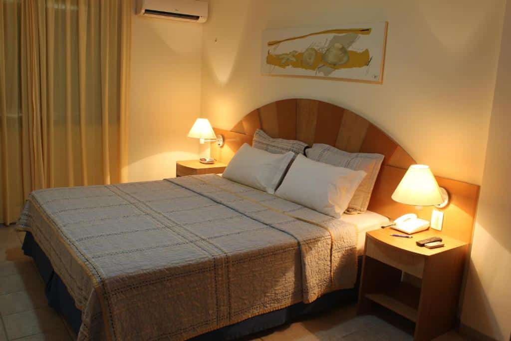 Uma cama de casal bem arrumada com duas mesinhas de cabeceira, abajures e um quadro acima da cabeceira em um quarto suavemente iluminado.