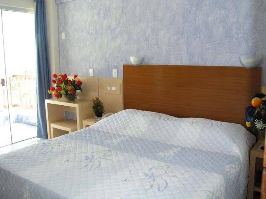 Uma cama bem arrumada com cabeceira de madeira, mesas laterais com flores e uma varanda visível por uma porta de vidro.