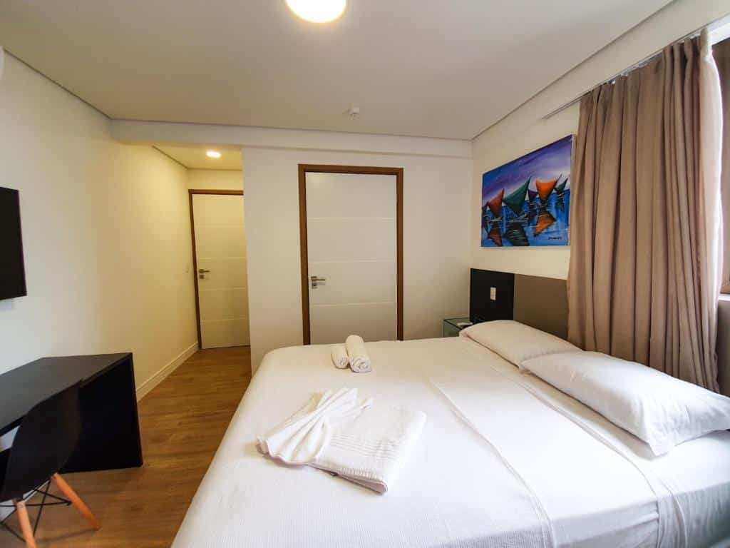 Um quarto de hotel moderno com uma cama de casal, uma escrivaninha com uma cadeira, toalhas dobradas e uma janela com cortinas bege.