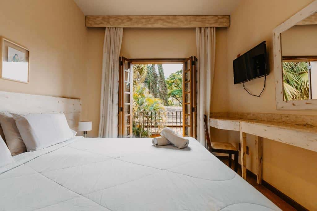 Quarto do Hotel Vila Bavária com uma varanda, uma televisão, uma bancada com cadeira, um espelho, uma cama de casal e um abajur