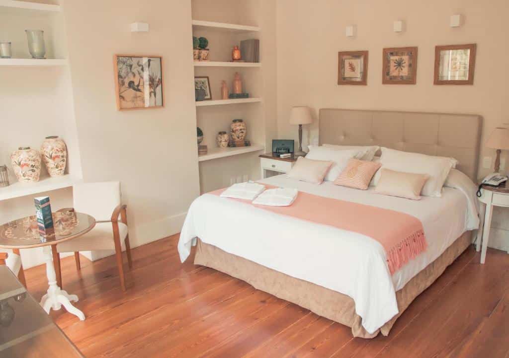 Quarto da Casa Rosa Hotel Boutique com chão de madeira, cama de casal, algumas estantes com enfeites, uma pequena mesa com duas cadeiras, e duas mesinhas de cabeceira com abajures