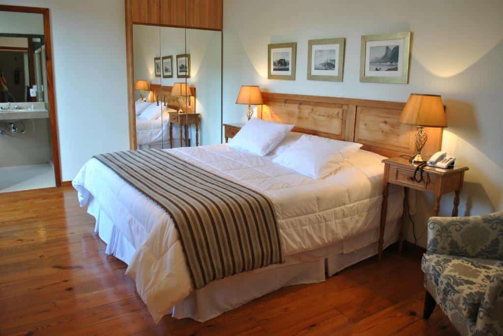 Quarto no Casablanca Koeler com cama de casal, chão de madeira, poltrona, duas mesinhas de cabeceira com abajur, um armário de duas portas com espelhos, para representar hotéis em Petrópolis