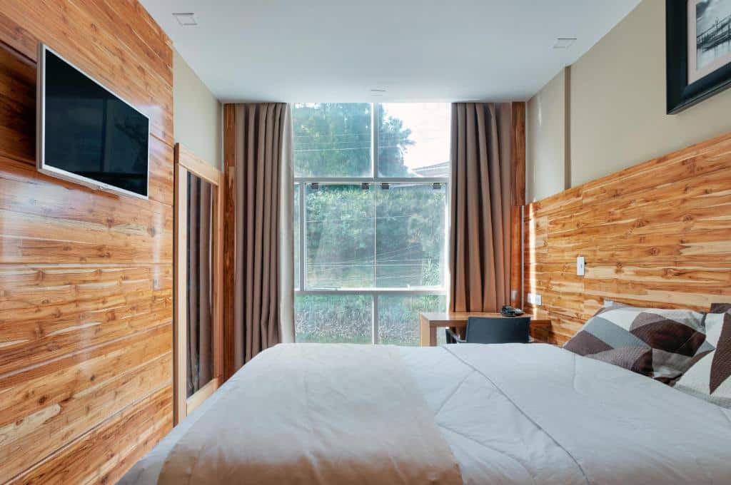 Quarto do Hotel Petropolis Inn com tudo decorado em madeira, há uma janela ampla com cortinas, um espelho de corpo inteiro, uma televisão, uma cama de casal e uma mesa de escritório com uma cadeira