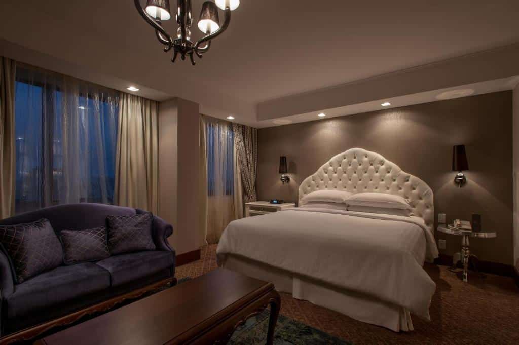 quarto do Sheraton com uma cama enorme de cabeceira suntuosa, além de um sofá e mesas de cabeceira e de centro