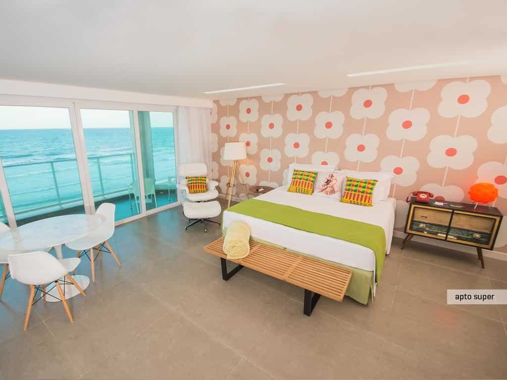 Um quarto bem iluminado do Kembali Hotel com uma cama grande, papel de parede retrô, vista para o mar e móveis modernos, incluindo uma pequena mesa e cadeiras.