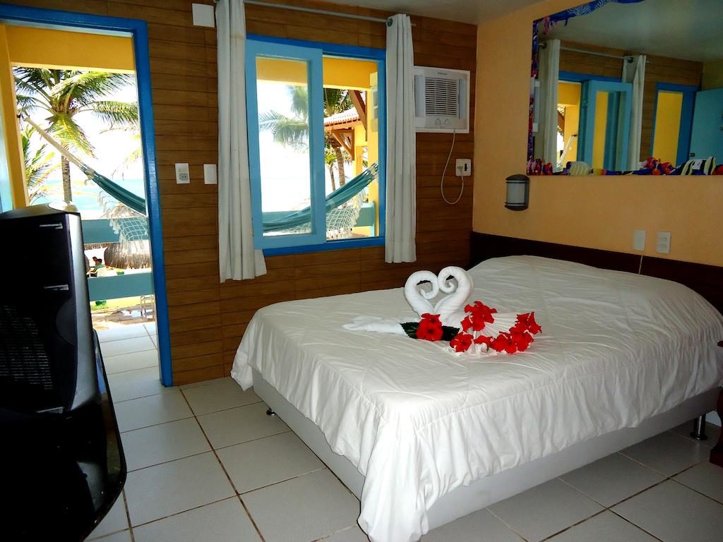 Quarto do Beira Mar Porto de Galinhas Hotel com uma cama de casal, uma toalha de cisne, TV, janelas, rede do lado de fora e palmeiras visíveis.