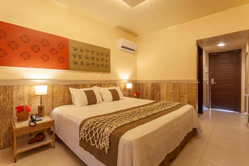 Um quarto do Ocaporã Hotel com uma cama grande, duas mesas de cabeceira com luminárias, arte na parede e uma porta que dá para o corredor.