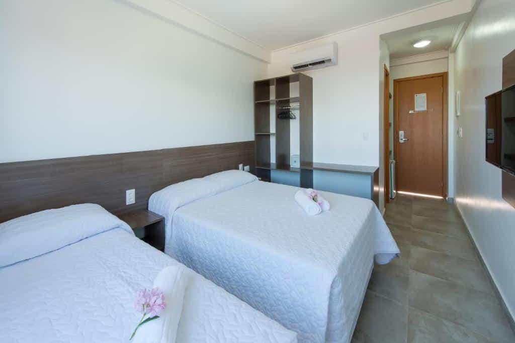 Um quarto do Hotel Vivenda dos Arrecifes com duas camas, roupa de cama branca, uma TV, armário aberto e uma porta de madeira; toalhas nas camas.
