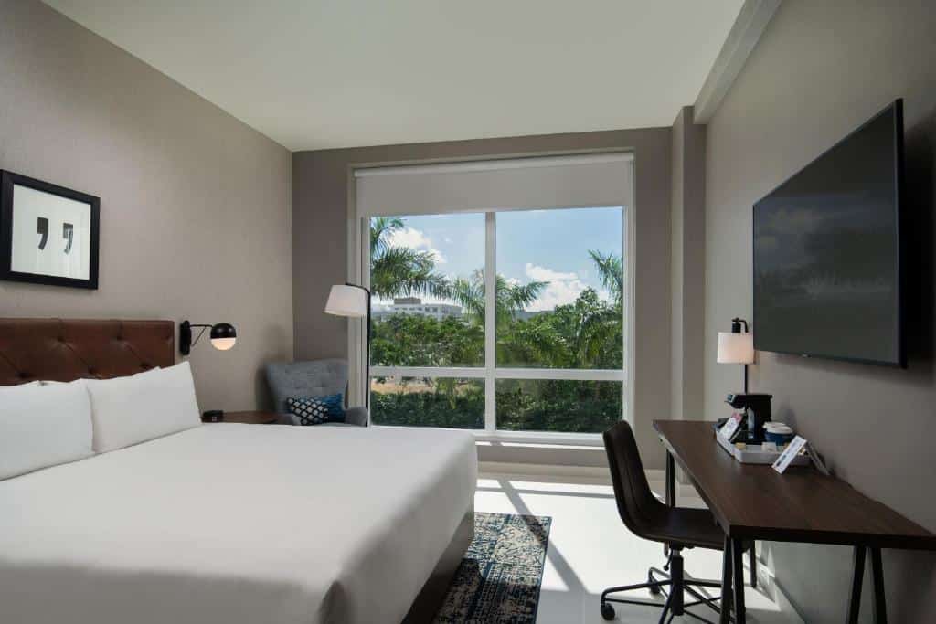 Quarto moderno do Four Points by Sheraton com uma cama grande, escrivaninha, poltrona e janela com vista para as palmeiras e o céu azul, para representar hotéis em Punta Cana.