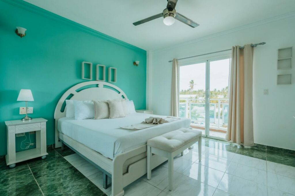 Quarto bem iluminado no Green Coast Hotel com uma parede com detalhes em azul-petróleo, cama branca, mesa de cabeceira, banco, ventilador de teto e portas de correr para uma varanda, para representar hotéis em punta Cana.
