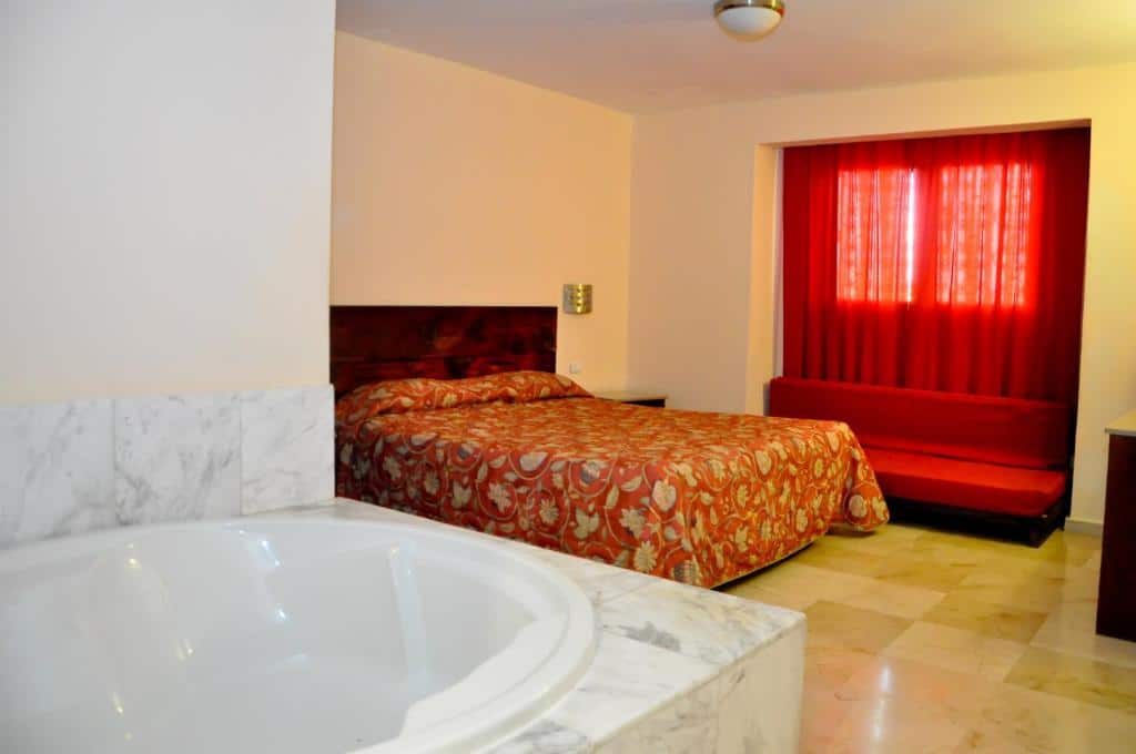 Quarto no Hotel Marimba Punta Cana com uma cama de casal, roupa de cama com estampa vermelha, um sofá vermelho, banheira de mármore e uma janela com cortinas vermelhas, para representar hotéis em Punta Cana.