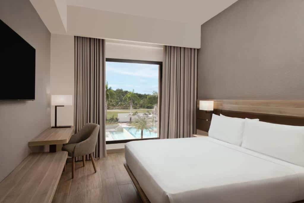 Quarto do AC Hotel by Marriott Punta Cana, um dos hotéis em Punta Cana. Uma cama de casal com mesinhas de cabeceira e abajures dos dois lados está encostada na parede do lado direito e encara uma longa mesa de trabalho com cadeira e abajur que tem uma TV na parede acima. Do lado direito da cama há uma janela com cortinas e vista para a piscina do hotel.