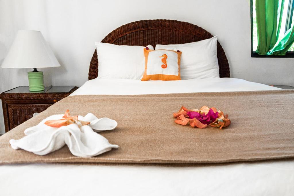Uma cama bem arrumada com um travesseiro de cavalo-marinho, uma manta marrom, uma toalha artística e pétalas de flores; abajur e janela de cada lado, no Riviera Punta Cana Eco Travelers, para representar hotéis em Punta Cana.