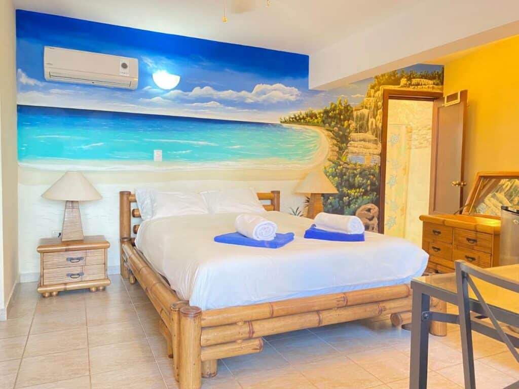 Quarto do Stanza Mare Beach Front, um dos hotéis em Punta Cana. Uma cama de bambu está encostada na parede central que tem estampa de praia e um aparelho de ar-condicionado, e conta com mesinhas de cabeceira e abajures dos dois lados. Do lado direito, uma cômoda fica em frente à porta que leva ao banheiro.
