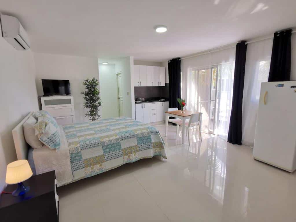 Quarto das SOL CARIBE SUITES, um dos hotéis em Punta Cana. Uma cama de casal tem mesinha de cabeceira com abajur do lado direito e uma cômoda com televisão do lado esquerdo, que fica ao lado de um vaso de planta. No fundo há uma área com cozinha e mesa com cadeiras. A parede em frente à cama tem janelas com cortinas e, do lado direito, uma geladeira.