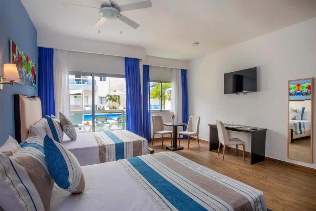 Quarto do whala!urban punta cana com duas camas, uma escrivaninha, uma TV na parede, uma pequena mesa com cadeiras e uma janela com vista para a área da piscina, para representar hotéis em Punta Cana.