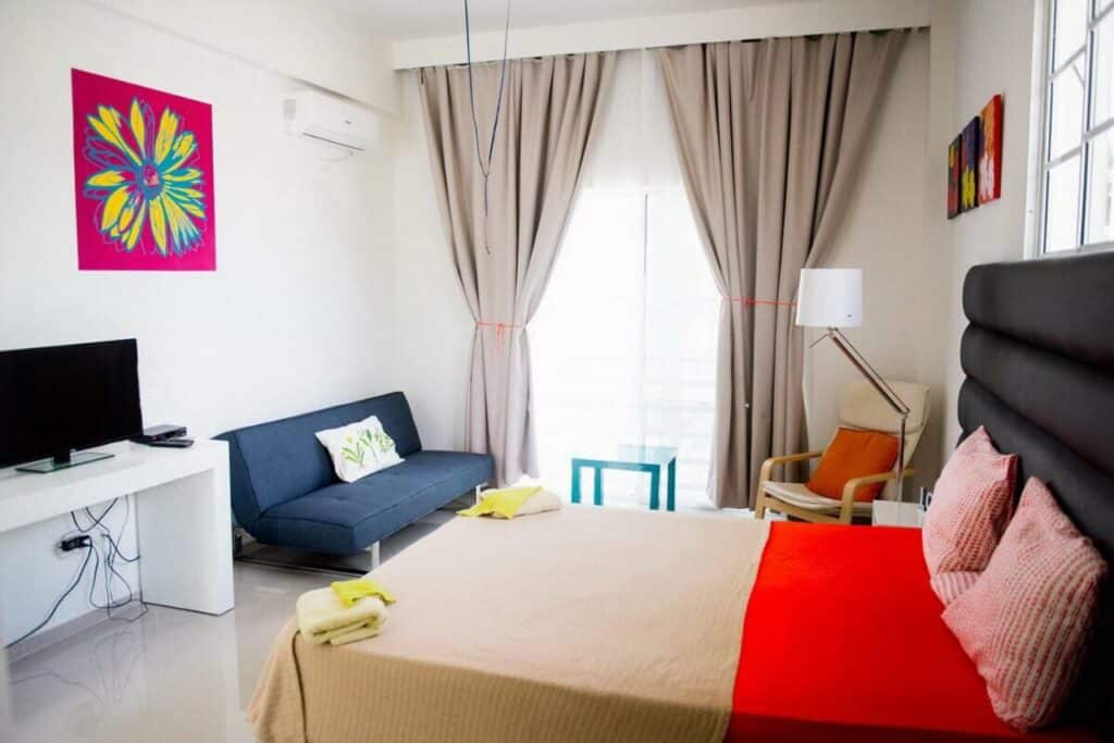 Um quarto moderno no Art Villa Dominicana com uma cama de casal, um sofá preto, TV, arte colorida na parede e uma grande janela com cortinas que deixa entrar a luz, para representar hotéis em Punta Cana.