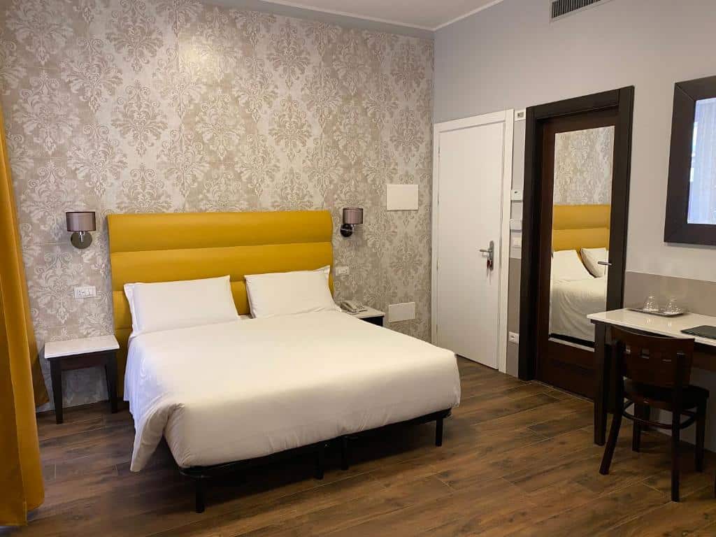 Quarto do Hotel Virgilio com uma cama de casal, cabeceira amarela, armário espelhado, escrivaninha com cadeira e papel de parede estampado.