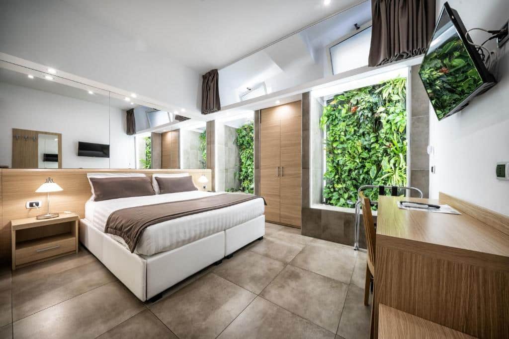 Quarto do Hotel Paolo II com cama de casal, TV na parede, escrivaninha e jardim interno vertical ao lado das portas de madeira do armário.
