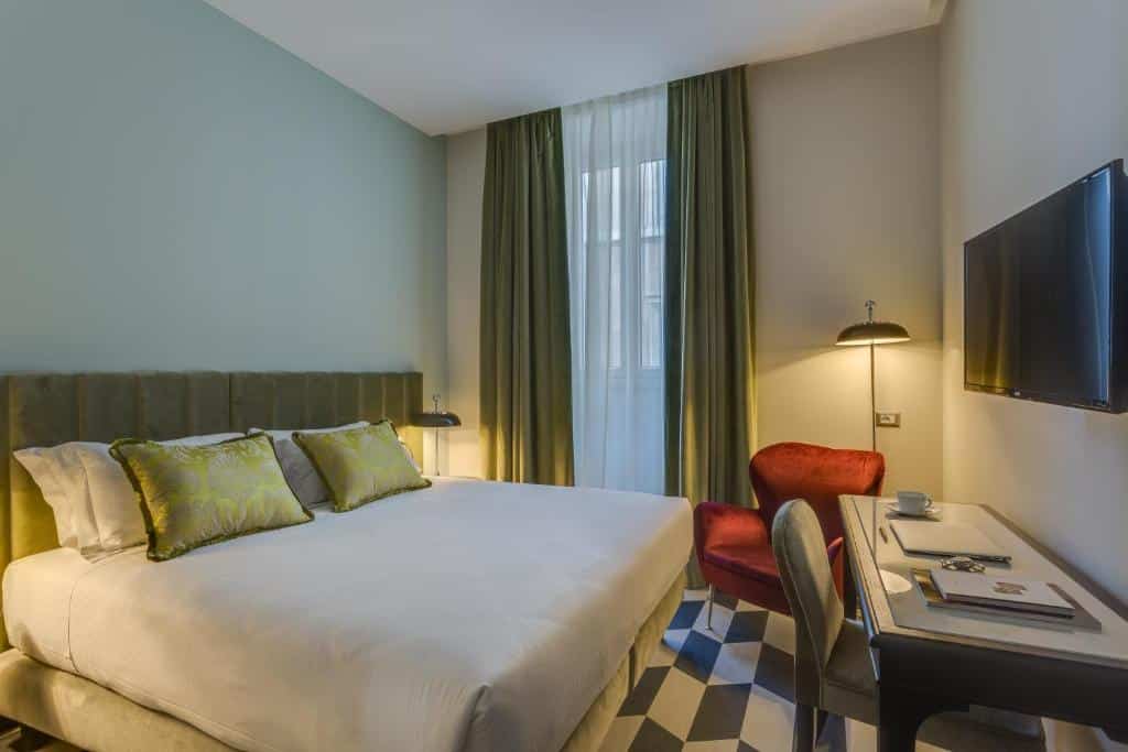 Um quarto do Otivm Hotel com uma cama de casal, travesseiros verdes, cadeira vermelha, escrivaninha, TV na parede e uma grande janela com cortinas verdes.