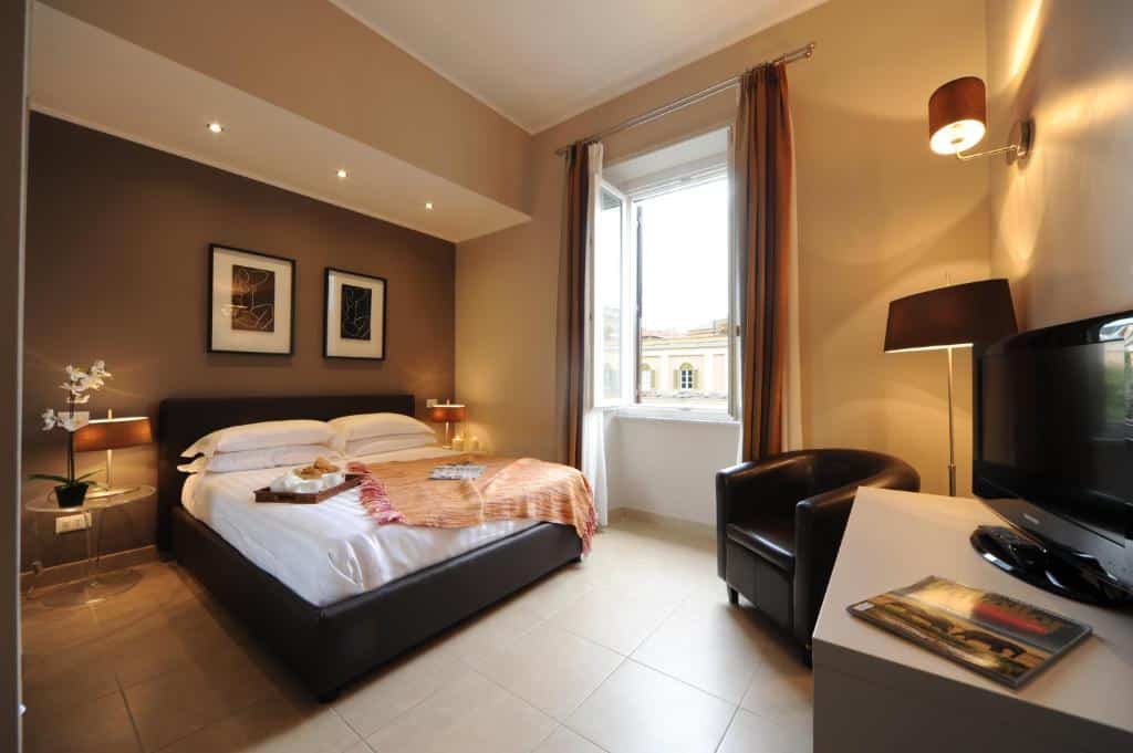 Quarto Trianon Borgo Pio Aparthotel com uma cama de casal, escrivaninha, poltrona, TV, janela com cortinas e decoração em tons neutros.