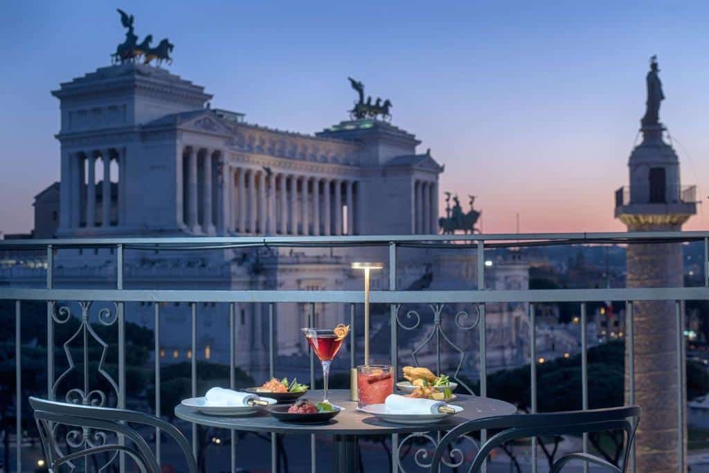 vista uma das opções de hotéis em Roma Uma mesa com alimentos e bebidas em uma varanda do NH Collection Roma Fori Imperiali, com vista para edifícios históricos e estátuas ao anoitecer. Este é um dos hotéis em Roma.