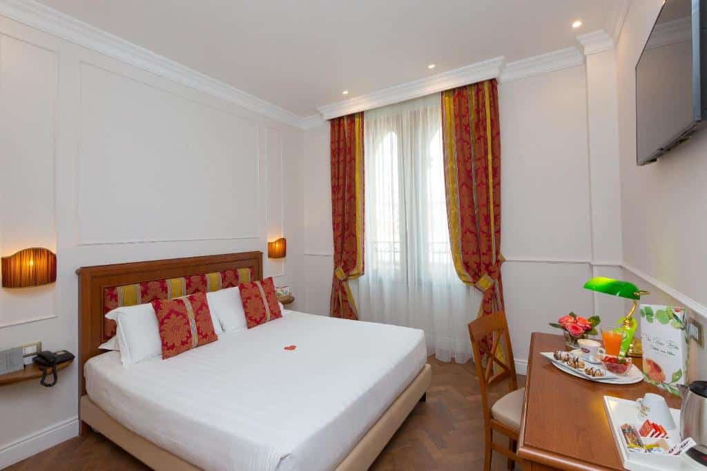 quarto do Hotel Milton Roma com uma cama de casal, escrivaninha, cadeira, luminárias de parede, TV e janela com cortinas vermelhas e douradas.
