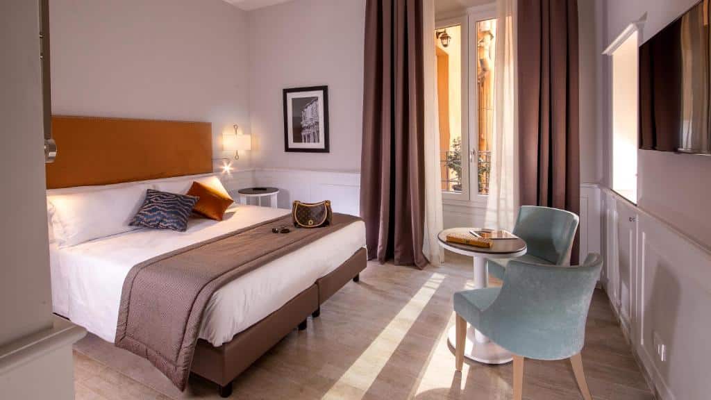 Um quarto do Princeps Boutique Hotel com uma cama de casal, duas cadeiras em uma mesa redonda, uma TV, uma janela grande com cortinas e decoração neutra.