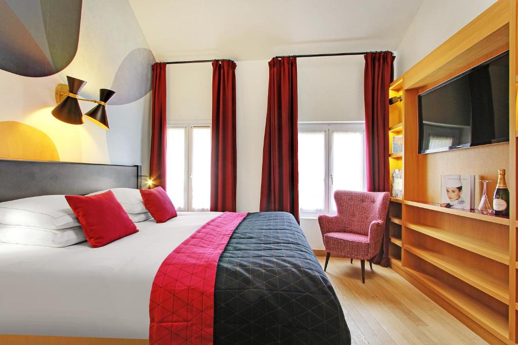 Quarto do Hôtel Artus, um dos hotéis em Saint Germain em Paris. Uma cama de casal está encostada na parede esquerda do local com mesinhas de cabeceira e abajures dos dois lados. Logo em frente fica uma estante com TV e garrafa de espumante com taças. Na lateral direita há duas janelas com cortinas na parede, e poltrona fica logo em frente a elas.