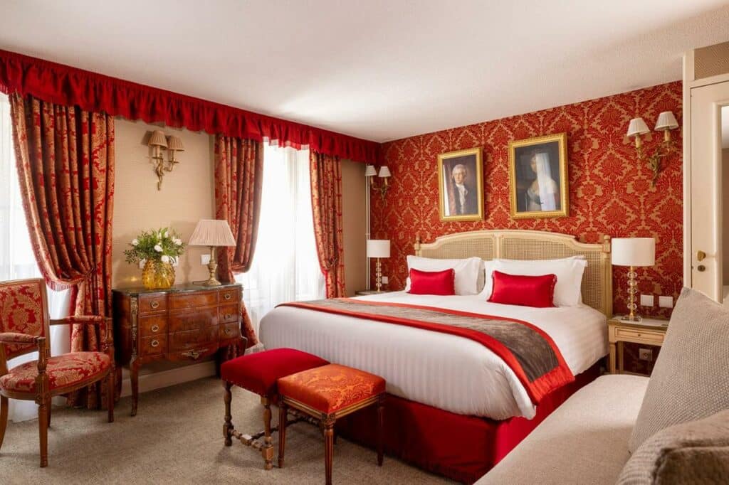 Quarto do Hotel De Seine, um dos hotéis em Saint Germain em Paris. Uma cama de casal está encostada na parede direita do local com mesinhas de cabeceira e abajures dos dois lados, assim como dois quadros na parede acima da cabeceira e banquinhos aos pés. Logo em frente fica uma área de estar com poltrona e sofá. Na lateral esquerda há duas janelas com cortinas na parede, e um aparador com abajur e vaso de flores fica entre elas.